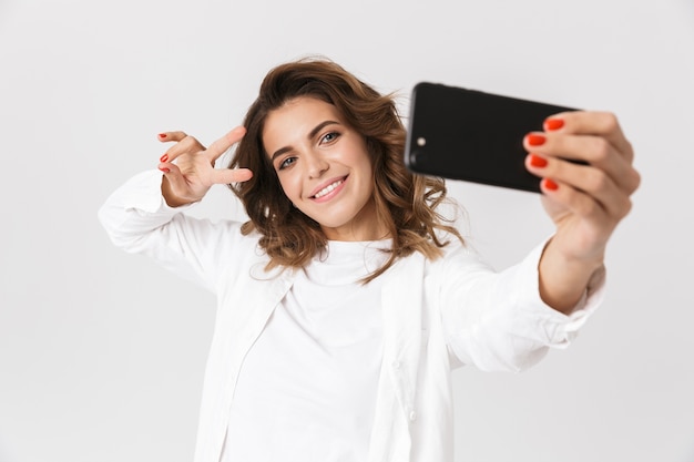 Portrait of attractive woman in casual clothes taking selfie photo on cell phone, while standing isolated