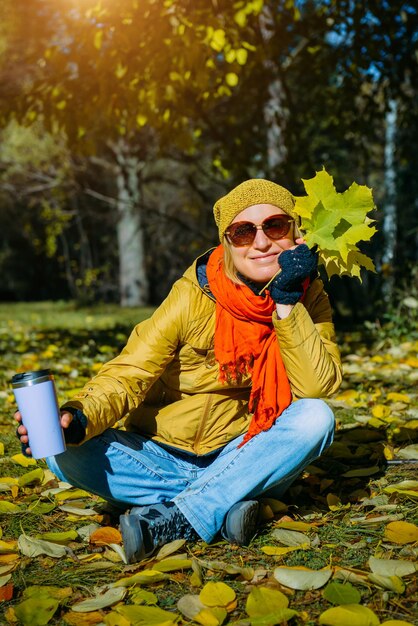 Portrait of attractive woman on a walk in autumn park sitting on yellow foliage in sunlight. Pretty girl holding maple leaves and thermo mug with a hot drink. Happy healthy lifestyle.