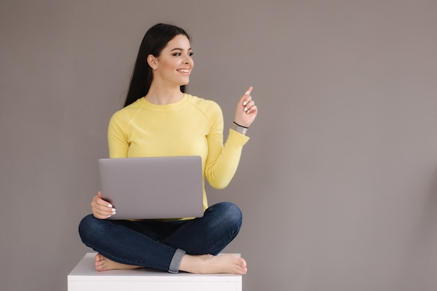 Portrait of attractive woman in yellow sweatshirt sitting with paltop and point the finger at place for text.