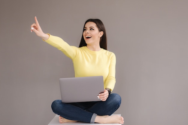 Portrait of attractive woman in yellow sweatshirt sitting with paltop and point the finger at place for text.