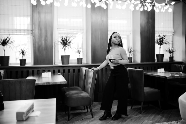 Portrait of attractive young african american woman wearing in green sweater and black jeans pose at restaurant with glass of red wine at hand