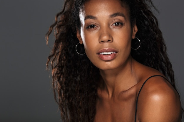 Portrait of an attractive young african woman with long curly hair posing while standing isolated over gray wall, looking 