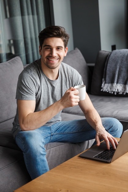 Portrait of an attractive young bearded man wearing casual clothes sitting on a couch at the living room, drinking coffee while using laptop computer