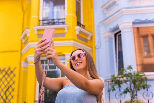 Portrait attractive young blonde woman taking a selfie behind yellow and blue colorful facade