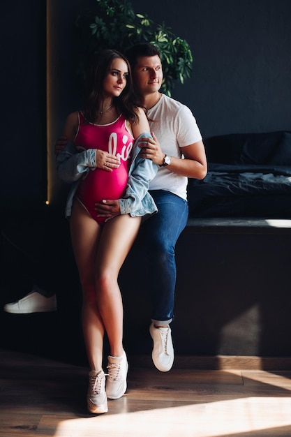 Portrait of attractive young couple sitting together Beautiful pregnant woman and handsome man hugging and holding hands together on female belly Happy family in casual clothes posing at camera