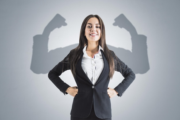 Portrait of attractive young european businesswoman with hands on sides and smile shadow muscle arms on concrete wall background Strenght and leadership concept