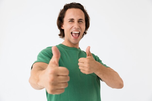 Photo portrait of attractive young man wearing basic t-shirt laughing and showing thumbs up isolated over white wall