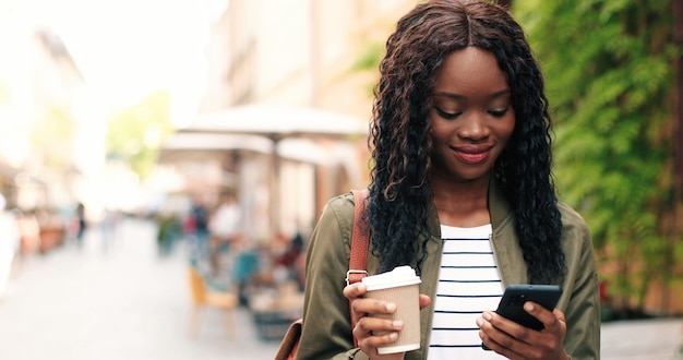 Portrait of the attractive young multiracial woman talking smartphone and drinking coffee outdoors b...