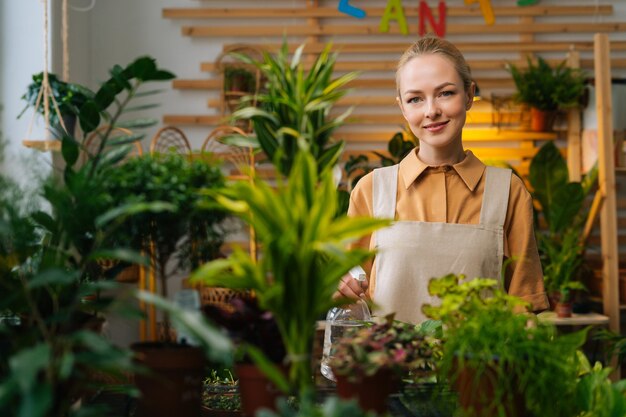 Portrait of attractive young woman in apron spraying water on houseplants in flower pots by sprayer looking at camera