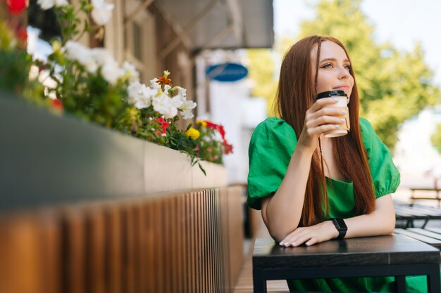 Photo portrait of attractive young woman enjoying tasty cup of coffee sitting at table in outdoor cafe terrace in summer day. pretty redhead lady relaxing leisure activity on urban street.