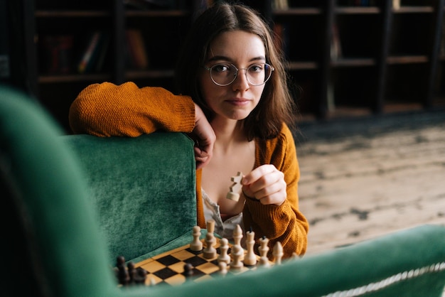 Portrait of attractive young woman wearing elegant eyeglasses making chess move with knight piece sitting in floor looking at camera