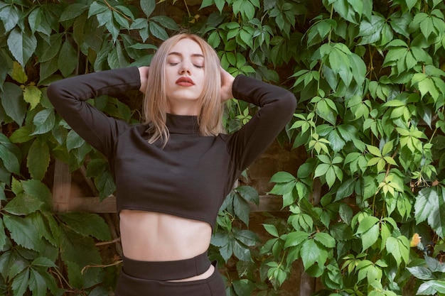 Portrait of attractive young woman with closed eyes blonde hair in short black top on green plants background