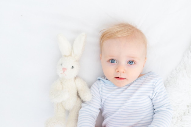 Portrait of a baby blonde boy six months old on a bed with a toy