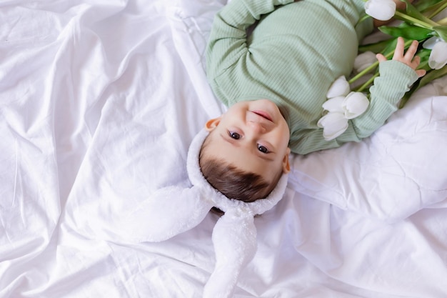 Portrait of a baby in a green cotton bodysuit with white rabbit ears lying on bed among white tulips