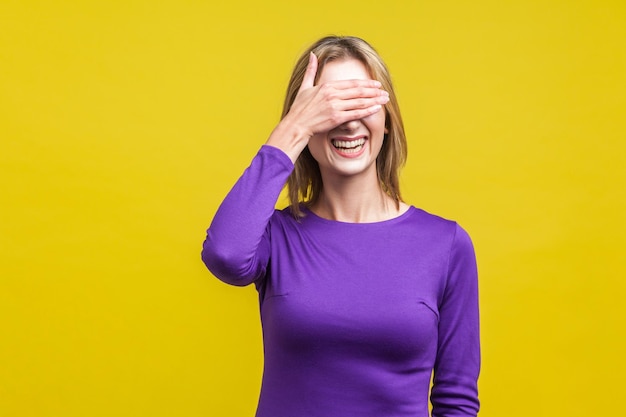 Portrait of bashful positive woman in elegant purple dress covering eyes with hand and laughing shy to look indoor studio shot isolated on yellow background