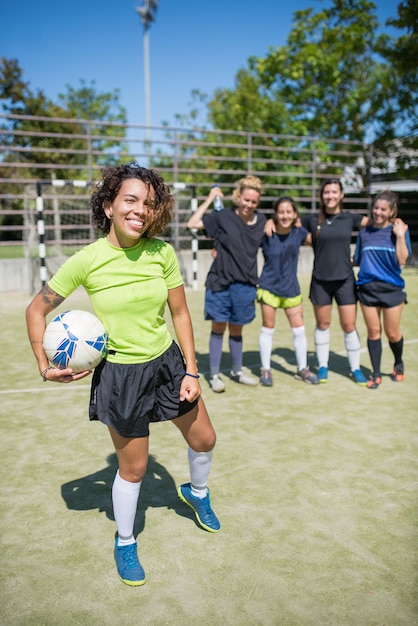 Portrait of beaming young woman on football field. Sportswoman with curly hair in uniform looking at camera, holding ball, cheerful teammates in background. Sport, leisure, active lifestyle concept