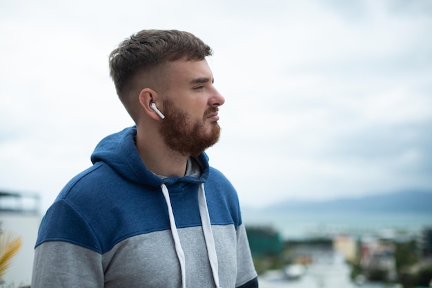 Portrait of bearded handsome pensive thoughtful guy listen music in earphones young upset sad frustrated man on natural mountain background thinking with a serious face