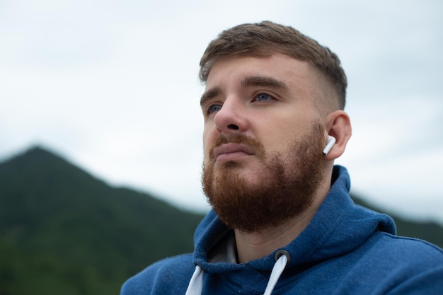 Portrait of bearded handsome pensive thoughtful guy listen music in earphones young upset sad frustrated man on natural mountain background thinking with a serious face