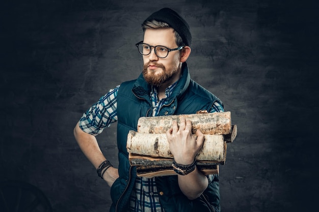 Portrait of a bearded male dressed in fleece shirt and warm waistcoat holds firewoods in his arms.