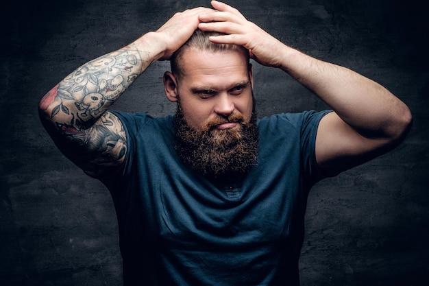 Portrait of bearded male touches his hairstyle with tattooed arms.