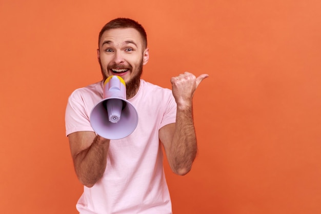 Portrait of bearded man holding megaphone and pointing aside, showing copyspace for advertisement with happy expression, wearing pink T-shirt. Indoor studio shot isolated on orange background.