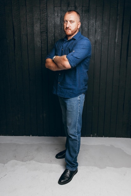 Portrait of a bearded man with a serious look standing on a black wooden background a business man in a blue shirt and jeans