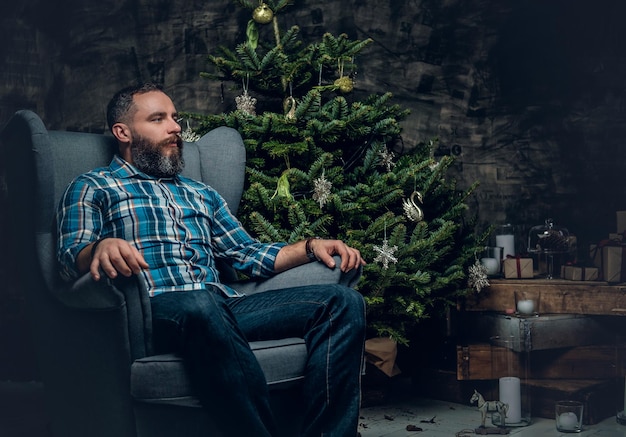 Portrait of bearded middle age male dressed in plaid flannel shirt and jeans sits on a chair over Christmas decorated background.