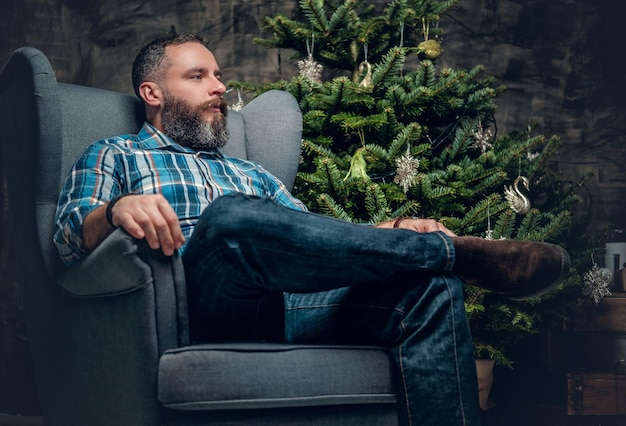 Portrait of bearded middle age male dressed in plaid flannel shirt and jeans sits on a chair over Christmas decorated background.