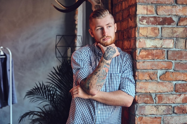 Portrait of a bearded modern male with tattoos on his arms posing near the window in a room with loft interior.