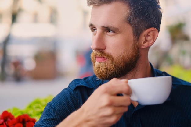 Portrait of bearded redhead casual man drinks coffee in a cafe on a street.