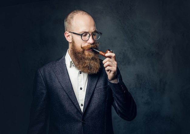 Portrait of bearded redhead English male smoking pipe over grey background.