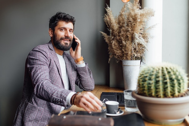 Portrait of bearded stylish man using laptop at cafe while sitting the wooden table, drinking coffee and speak by phone