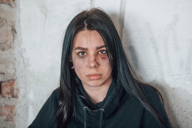 Portrait of beaten young woman with bruise under eye that standing and leaning on the wall indoors in abandoned building