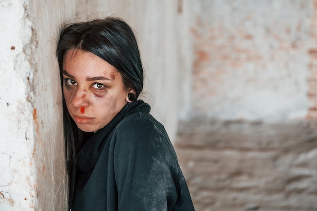 Portrait of beaten young woman with bruise under eye that standing and leaning on the wall indoors in abandoned building