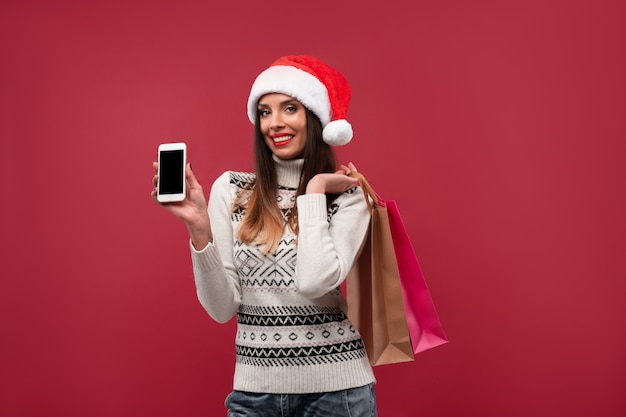 Portrait beautifiul caucasian woman Santa hat on red  wall. Christmas New Year  concept. Cute woman teeth smiling positive emotions with shopping bag and phone Online shopping