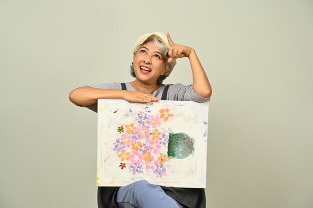 Portrait of beautiful 60s senior woman wearing apron holding canvas and pointing finger overhead isolated on gray background