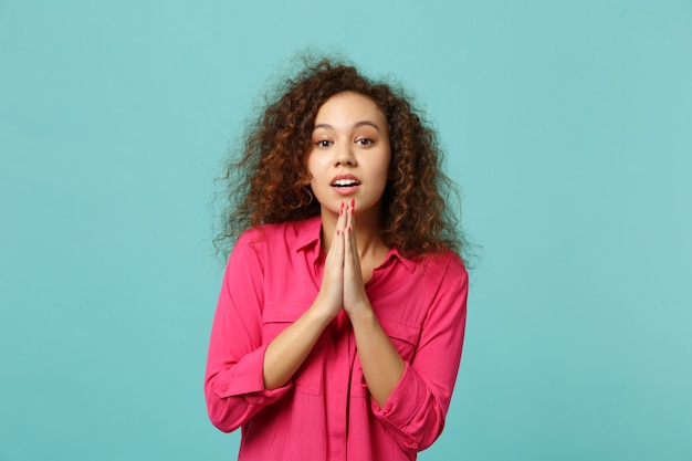 Portrait of beautiful african girl in casual clothes folded his hands and praying isolated on blue turquoise wall background in studio. People sincere emotions, lifestyle concept. Mock up copy space.