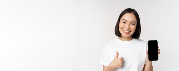 Portrait of beautiful asian woman showing thumb up and smartphone screen smiling while advertising m