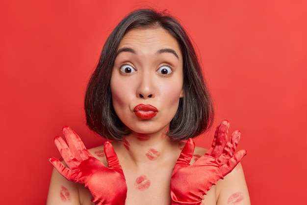 Portrait of beautiful Asian woman with dark hair red painted lips looks surprisingly at camera raises hands in gloves has glamour look posess shortless with kiss traces on body isolated over red wall