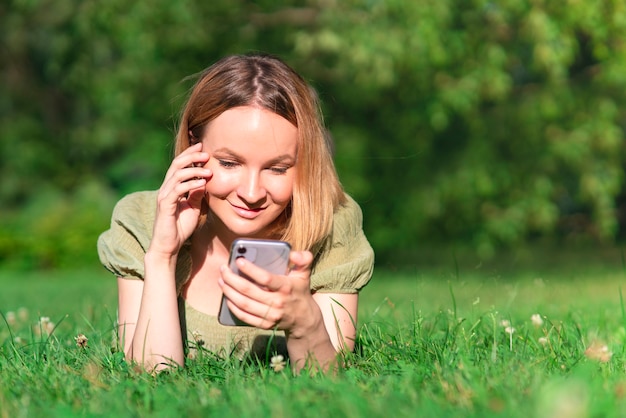 Portrait of beautiful attractive girl, young happy cheerful positive woman is lying in summer park on grass, smiling and looking at screen of her cell mobile phone, reading message, social media