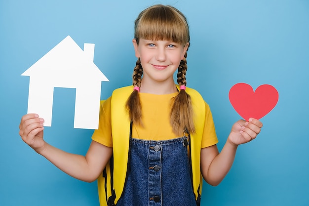 Portrait of beautiful attractive smiling school girl holding small red heart and paper white house model, happy looking at camera, wears yellow backpack, isolated over blue color background in studio