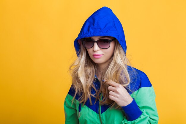 Portrait of beautiful blond woman in sunglasses and blue green hooded jacket on yellow background hi...