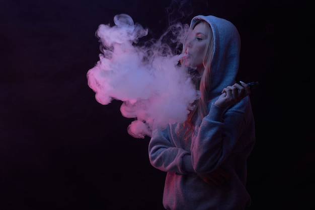 Photo portrait of beautiful blonde girl in gray hoodie smokes vape isolated on black studio background, cloud of steam smoke, mini hookah