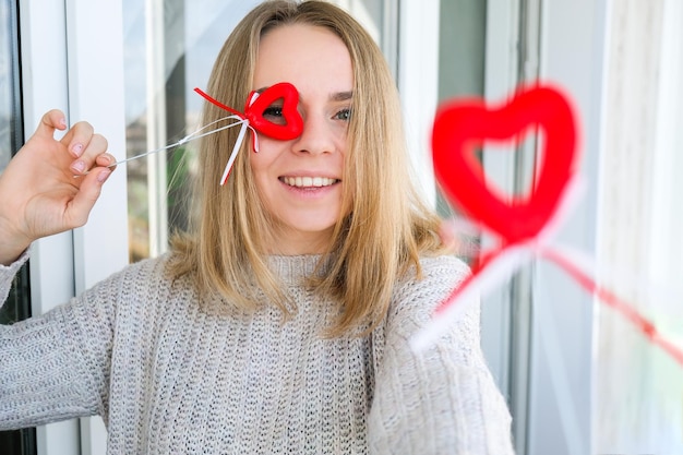 Portrait of beautiful blonde happy woman holding symbol heart. Two red hearts. Girl taking selfie. Alone on Valentines day. Love. Romantic. White sweater, emotions