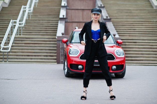 Portrait of beautiful blonde sexy fashion woman model in cap and in all black blue corset with bright makeup near red city car