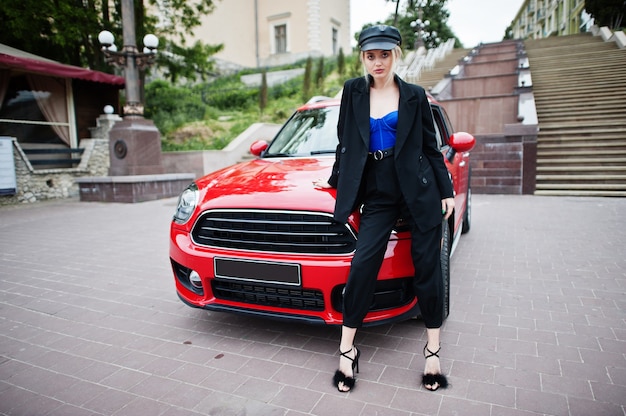 Portrait of beautiful blonde sexy fashion woman model in cap and in all black, blue corset, with bright makeup near red city car.