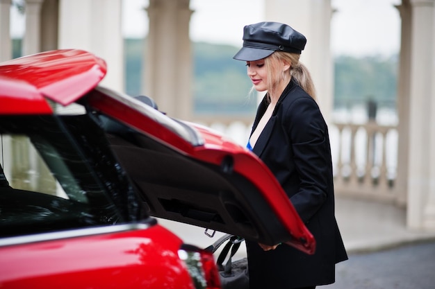 Portrait of beautiful blonde sexy fashion woman model in cap and in all black with bright makeup near red city car put her bag in trunk