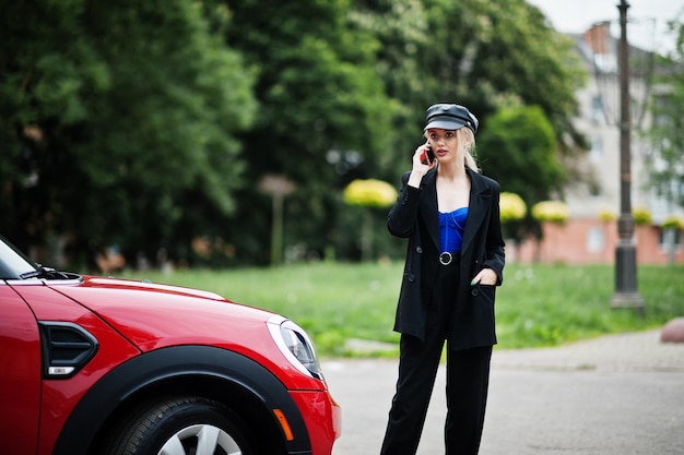 Portrait of beautiful blonde sexy fashion woman model in cap and in all black with bright makeup near red city car with mobile phone