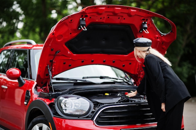 Portrait of beautiful blonde sexy fashion woman model in cap and in all black with bright makeup near red city car with open hood