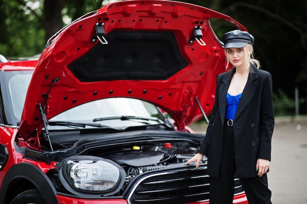 Portrait of beautiful blonde sexy fashion woman model in cap and in all black with bright makeup near red city car with open hood